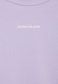 Světle fialové bavlněné tričko s kulatým výstřihem, žebrovanou strukturou a bílým logem "CALVIN KLEIN JEANS" uprostřed hrudi.