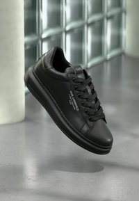 Fekete bőr sneaker sima felülettel, kerek orral, texturált díszítésekkel a sarokrészen és fekete fűzőkkel. Az oldalon logódiszítés található.