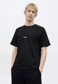 Fekete pamut rövid ujjú póló kerek nyakkivágással és fehér "JJ Studios" logóval a mellkason, fekete nadrággal párosítva.