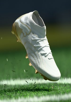 Balta futbola zābaku ar zelta akcentiem sitot pa zāli zaļā laukumā ar baltas līnijas atzīmi.