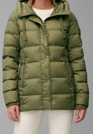 Žena nosící olivově zelenou prošívanou péřovou bundu s kapucí, zapínáním na zip a patentky vpředu a zipovými kapsami na bocích, doplněnou bílými kalhotami.