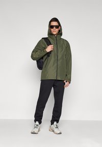 Grüne wasserdichte Jacke mit Kapuze, kombiniert mit schwarzen Jogginghosen und grauen Sportschuhen. Das Modell trägt einen schwarzen Rucksack und eine Sonnenbrille.