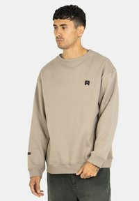 Beiger Sweatshirt mit Rundhalsausschnitt, gerippten Bündchen und einem kleinen schwarzen Logo auf der Brust. Weicher, strukturierter Stoff mit einer lockeren Passform.