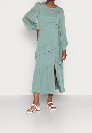 Teal midi wrap-kjole med flæser, gennemsigtige biskopærmer, bundet talje, frontslids, båret med hvide sandaler med blokhæl og stropper.