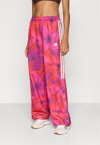 Ροζ παντελόνι φαρδιάς γραμμής με λουλουδάτο μοτίβο και ελαστική μέση, με λευκές ρίγες Adidas και κουμπιά στο πλάι.