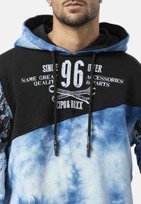 Sweatshirt met capuchon met een zwart bovenstuk, een blauw tie-dye onderstuk en contrasterende witte tekst en afbeeldingen. Inclusief verstelbare trekkoorden.