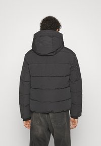 Jachetă neagră cu umplutură, cu glugă, având secțiuni orizontale quilted, manșete elastice și o croială lejeră. Vedere din spate.