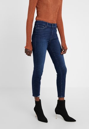 Woman wears dark blue skinny jeans, burnt orange ribbed long-sleeve top, and black pointed-toe high-heeled ankle boots on plain background.