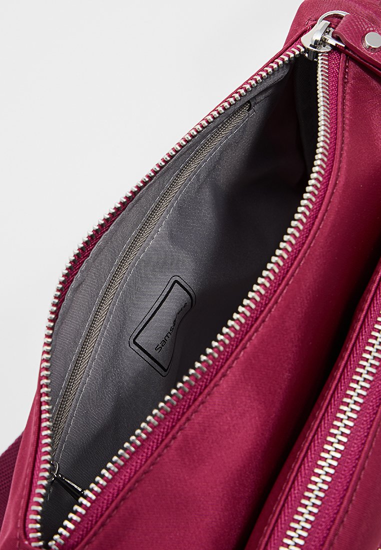 Bolso de tela burdeos con un forro interior suave y cremallera plateada. Presenta una pequeña etiqueta y un amplio compartimento principal. La cremallera se extiende completamente.