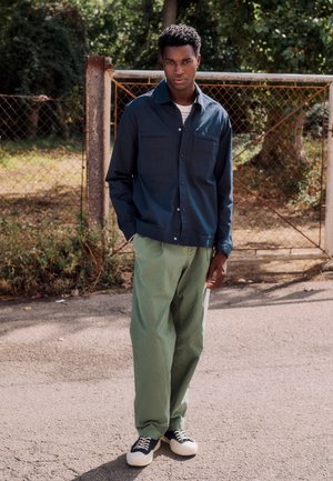 Mladi moški stoji na prostem blizu kovinske ograje, oblečen v mornarsko modro jakno, svetlo zelene hlače in črno-bele superge.