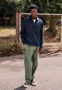 Ung mann som står utendørs ved en metallport, iført en marineblå jakke, lysegrønne bukser og svarte og hvite joggesko.