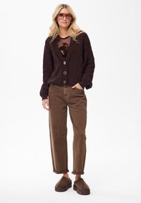 Brązowy sweter z dzianiny z dekoltem w serek i zapięciem na guziki, w połączeniu z brązowymi spodniami z wysokim stanem i brązowymi butami wsuwanymi. Jasne dodatki.