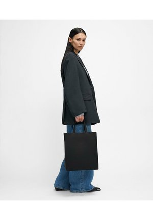 Borsa tote in pelle nera dalla forma quadrata, con manici corti e texture liscia, tenuta da una persona che indossa un blazer grigio oversize e jeans di denim.