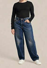 Personne portant un jean large bleu foncé, une chemise noire à manches longues, des baskets blanches, debout avec une main dans la poche sur un fond uni.