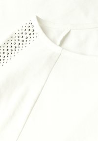 Camiseta blanca de cuello redondo con adornos de tachuelas plateadas en un hombro. Textura suave de algodón, diseño casual.