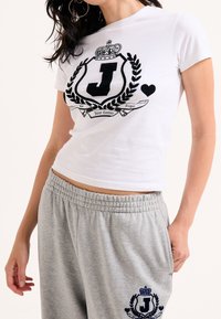 T-shirt bianco a maniche corte con una grafica nera che presenta una "J" e una corona, abbinato a pantaloni sweatpants grigi con vita elastica e un logo.