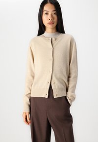 Beige kardigan pehmest kangast, ümmarguse kaelusega, nööpide sulgemisega ja soonikute vaheliste mansettidega. Kandmisest pruunide pükste kohal.