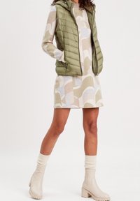 Grøn polstret vest over en mønstret kjole med bløde nuancer af beige, creme og lyseblå. Cremefarvede knæhøje støvler og strikkede sokker fuldender looket.
