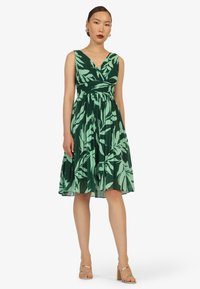 Grünes und blassgrünes, blumenmusterndes Kleid mit V-Ausschnitt, plissiertem Rock und figurbetontem Oberteil; kombiniert mit nude-farbenen Riemchenhemdchen.