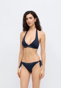 Σετ μπικίνι σε ναυτικό μπλε χρώμα με τριγωνικό τοπ και λουριά που δένουν γύρω από το λαιμό, καθώς και σλιπ χαμηλής μέσης με δεσίματα στα πλαϊνά και λογότυπο της μάρκας.