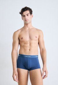 Borstvoeding man staat, draagt blauwe Calvin Klein boxer shorts, kijkt lichtjes naar beneden tegen een effen lichte achtergrond.