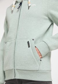 Svetle zelená mikina s kapucí na zip, s žebrovanými manžetami a dvěma předními kapsami. Obsahuje dekorační šňůrky s korálky a vyšitým logem.