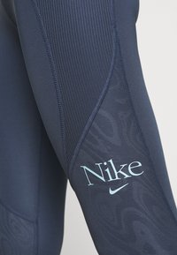 Námořnické sportovní legíny s ribbovanou texturou, hladkými panely a jemným vzorem. Na stehně se nachází světle modré logo Nike.
