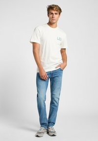 Camiseta de algodón de color claro con el logo "LEE" en azul, combinada con jeans azul claro y zapatillas grises. Ajuste simple y casual.