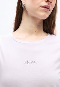 Γυναίκα που φοράει ένα ανοιχτό μωβ μπλουζάκι με στρογγυλή λαιμόκοψη, με ένα μικρό κεντητό λογότυπο "BOSS" κεντραρισμένο κοντά στη λαιμόκοψη και ασημένια κρίκους σκουλαρίκια.