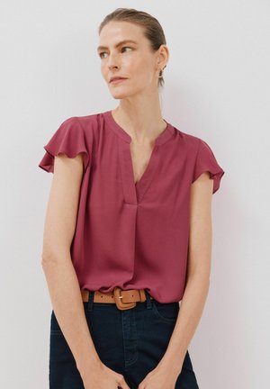 Kurzärmlige, tief burgunderrote Bluse mit V-Ausschnitt und subtilen Rüschensäumen, kombiniert mit einer dunklen Jeans und einem braunen Gürtel. Glatte Stoffoberfläche.