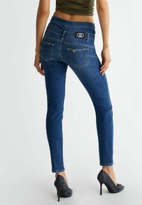 Jean skinny bleu avec une taille haute, deux poches arrière et un patch logo. Accompagné de chaussures noires à talons hauts ornées de strass.