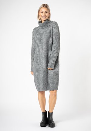 Graues, geripptes Rollkragenkleid mit langen Ärmeln, knielang, lockere Passform, mit einer weichen Textur und dezentem Saumfransen. Getragene schwarze Ankle Boots.