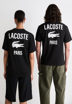 Du žmonės stovi nugaromis, vilkėdami juodus Lacoste Paris marškinėlį su baltais krokodilų logotipais, vienas derinamas su juodomis šortais, o kitas su žaliomis kelnėmis.
