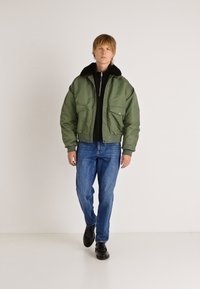 Cazadora bomber verde con cuello negro y bolsillos delanteros, combinada con vaqueros oscuros y zapatos negros. La modelo está de pie frente a un fondo liso.