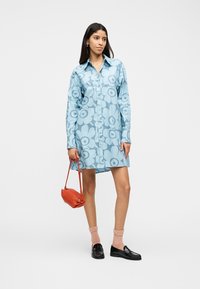 Γυναίκα που φοράει μπλε φόρεμα με λουλούδια και κουμπιά, κρατώντας μια μικρή κόκκινη τσάντα, σε συνδυασμό με ροζ κάλτσες και μαύρα μοκασίνια, στέκεται σε λευκό φόντο.