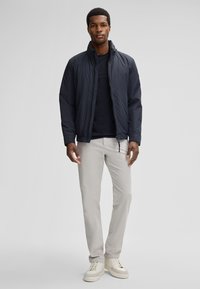 Giubbotto imbottito blu navy con colletto alto, chiusura con zip, indossato sopra una maglietta nera; pantaloni grigio chiaro e sneaker beige completano l'outfit.