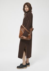 Strikket, ribbet brun kjole med sideslids; model holder en beige lædertaske med kædedetaljer; sorte sko med tykke såler.