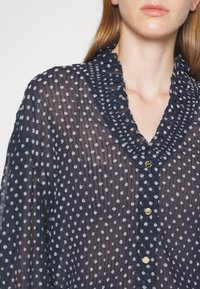 Gros plan d'une femme portant un chemisier transparent bleu marine à pois blancs, col en V à volants et boutons dorés sur le devant.