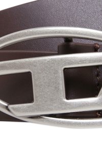 Brązowy skórzany pasek z metalową owalną klamrą o wyciętym wzorze i teksturowanej powierzchni, podkreślony kontrastującymi szczegółami ściegów.