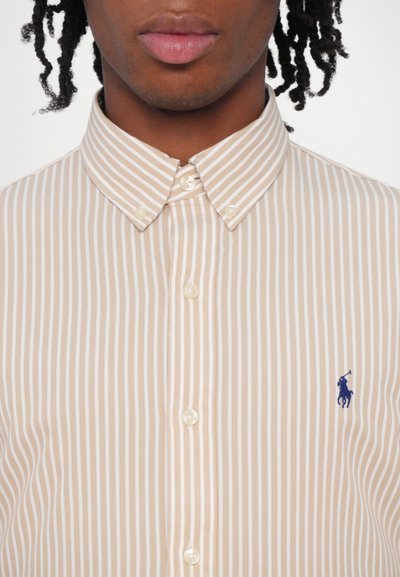 Šviesiai ruda ir balta juostele pabrėžta marškiniai su mygtukų užsegimu, puošti perlų mygtukais ir mėlynu siuvinėtų logotipu kairėje krūtinės pusėje.