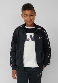Zwarte ritsjack met witte accenten en elastische manchetten, gecombineerd met een zwart-wit graphic t-shirt. Draagt een bijpassende zwarte broek.