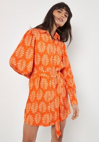 Oranges Hemd-Kleid mit Taillengürtel, versehen mit einem cremefarbenen Blättermuster und puffärmel, gefertigt aus leichtem Stoff.