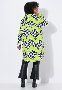 Donna con capelli ricci che indossa un long hoodie a zigzag e a vortice in verde neon, nero e bianco, pantaloni in pelle nera a campana e stivali neri.