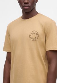 Homme portant un t-shirt marron clair avec un logo rond indiquant "Hugo Jeans" sur la poitrine gauche.