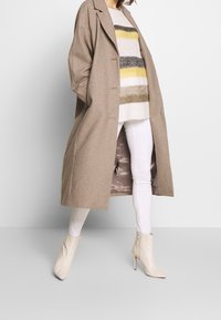 Cappotto di lana beige con una vestibilità rilassata, caratterizzato da un ampio revers e chiusura a bottone, indossato sopra un maglione a righe e jeans skinny bianchi, abbinato a stivaletti beige.