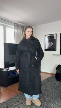 Cappotto lungo nero con una ampia colletto, bottoni doppi, vita con cintura, vestibilità oversize, indossato sopra jeans blu chiaro e scarpe beige.