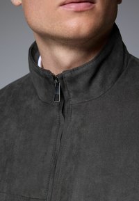 Veste en suède grise avec un col haut, fermeture zippée demi-longueur et texture lisse. Présente un léger éclat et des coutures soignées autour du col.