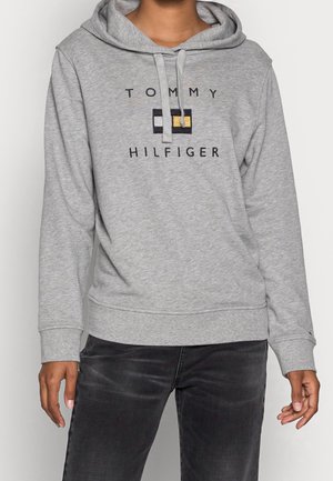 Personne portant un sweat à capuche gris clair Tommy Hilfiger avec cordon et un pantalon noir, debout devant un fond uni.