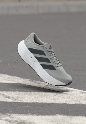 Zapatilla de correr gris claro con rayas negras, suela blanca, mostrada en el aire sobre una carretera de asfalto texturizada.