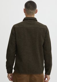 Camisa de lana marrón oscuro con una superficie texturizada, puños abotonados, cuello doblado y dobladillo recto. Presenta un detalle de yugo en la parte posterior.